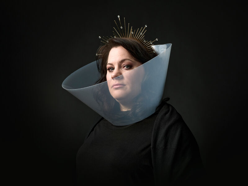 Berit Companjen draagt een brede, doorschijnende kraag en een gouden hoofdtooi met stralen tegen een donkere achtergrond.