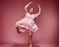 Persoon (Fimme Bakker) in roze tutu en balletpakje staat op spitzen met één been opgetild en armen gebogen boven het hoofd tegen een roze achtergrond.