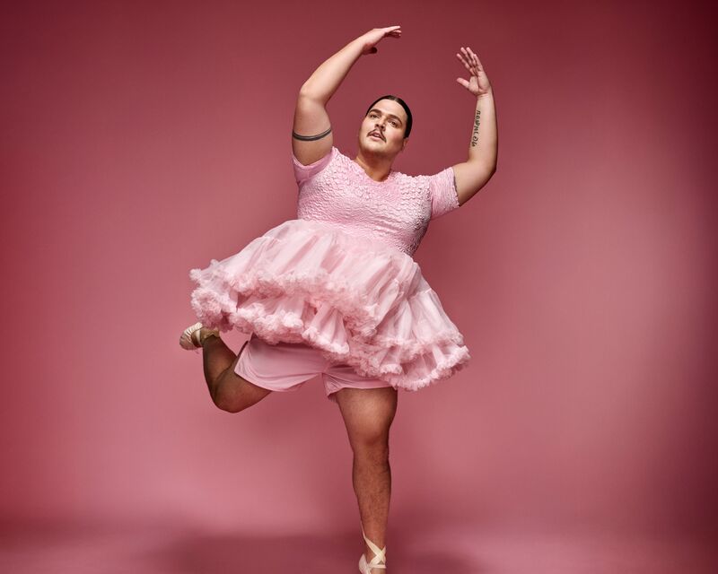 Persoon (Fimme Bakker) in roze tutu en balletpakje staat op spitzen met één been opgetild en armen gebogen boven het hoofd tegen een roze achtergrond.